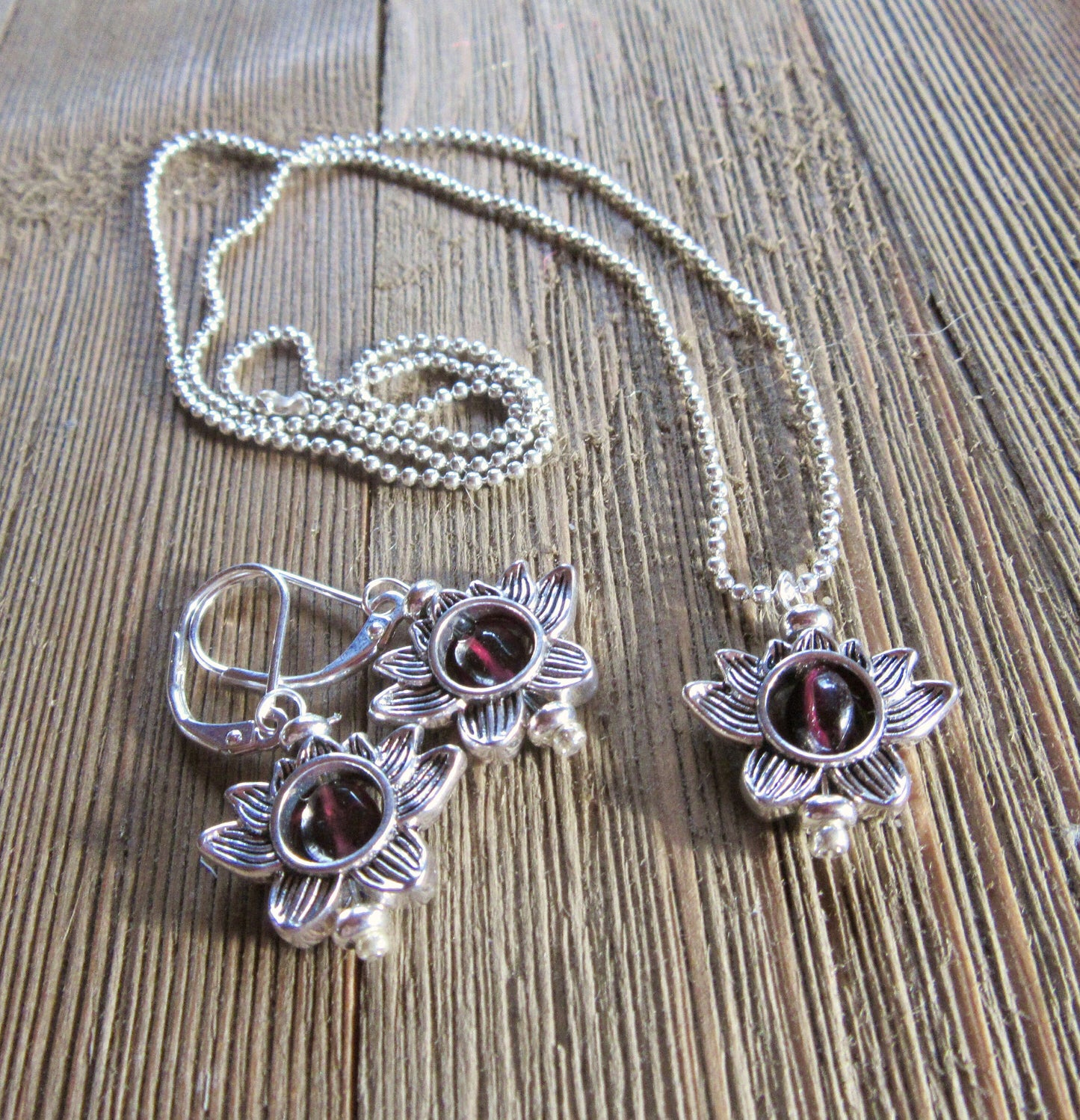 The Lotus Flower Zen Jewelry Set features silver lotus earrings, a dainty necklace with a garnet stone pendant, and a turquoise silver lotus charm, all beautifully displayed on a rustic wooden surface.