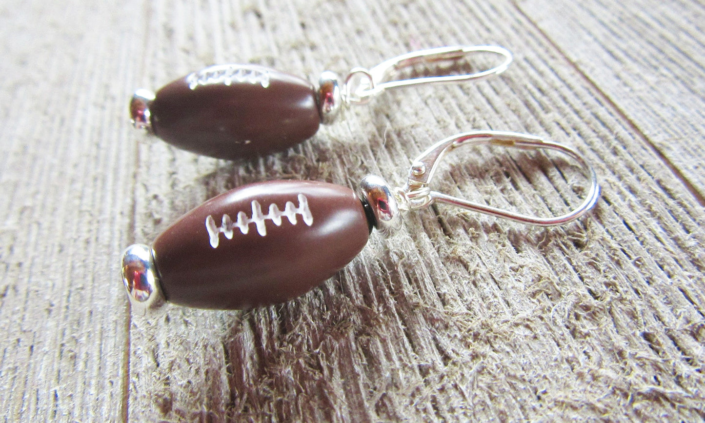 Football Earrings: Sterling Silver Sports Jewelry Gift for the big game, featuring small brown footballs with white stitching on sterling silver hooks, displayed on a textured wooden surface.