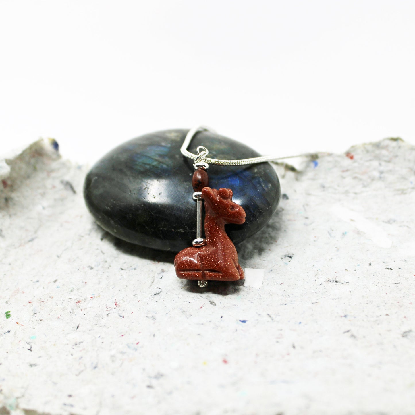 The Handcrafted Goldstone Giraffe Charm Pendant, designed for animal lovers, features a silver giraffe charm and goldstone pendant displayed on a textured gray surface with a polished dark stone in the background.
