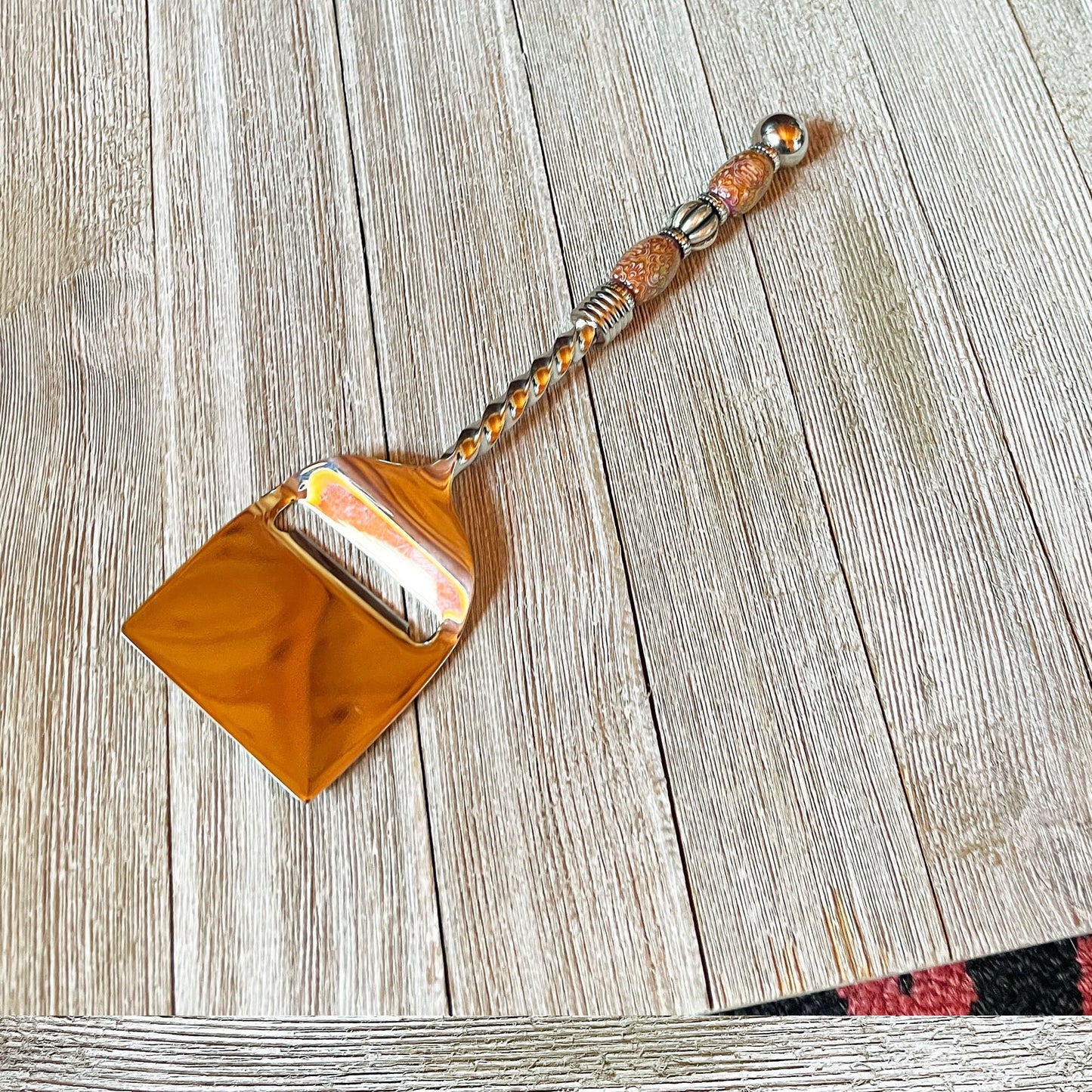 The Color Changing Beaded Cheese Slicer - Stainless Steel Party Tableware, featuring a decorative handle, sits on a light wooden surface that showcases its unique beaded design and the wood's visible grain patterns.
