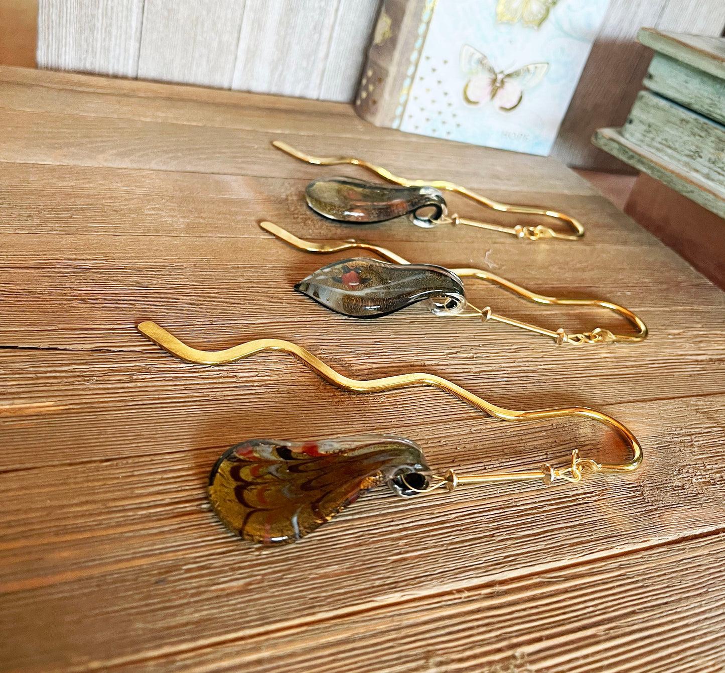 Three Art Glass Hook Bookmarks in gold and brown, each with a curved handle and leaf-shaped charm, rest on wood. Books blur in the background, highlighting this elegant reader gift: the Art Glass Hook Bookmark—Gold & Brown.