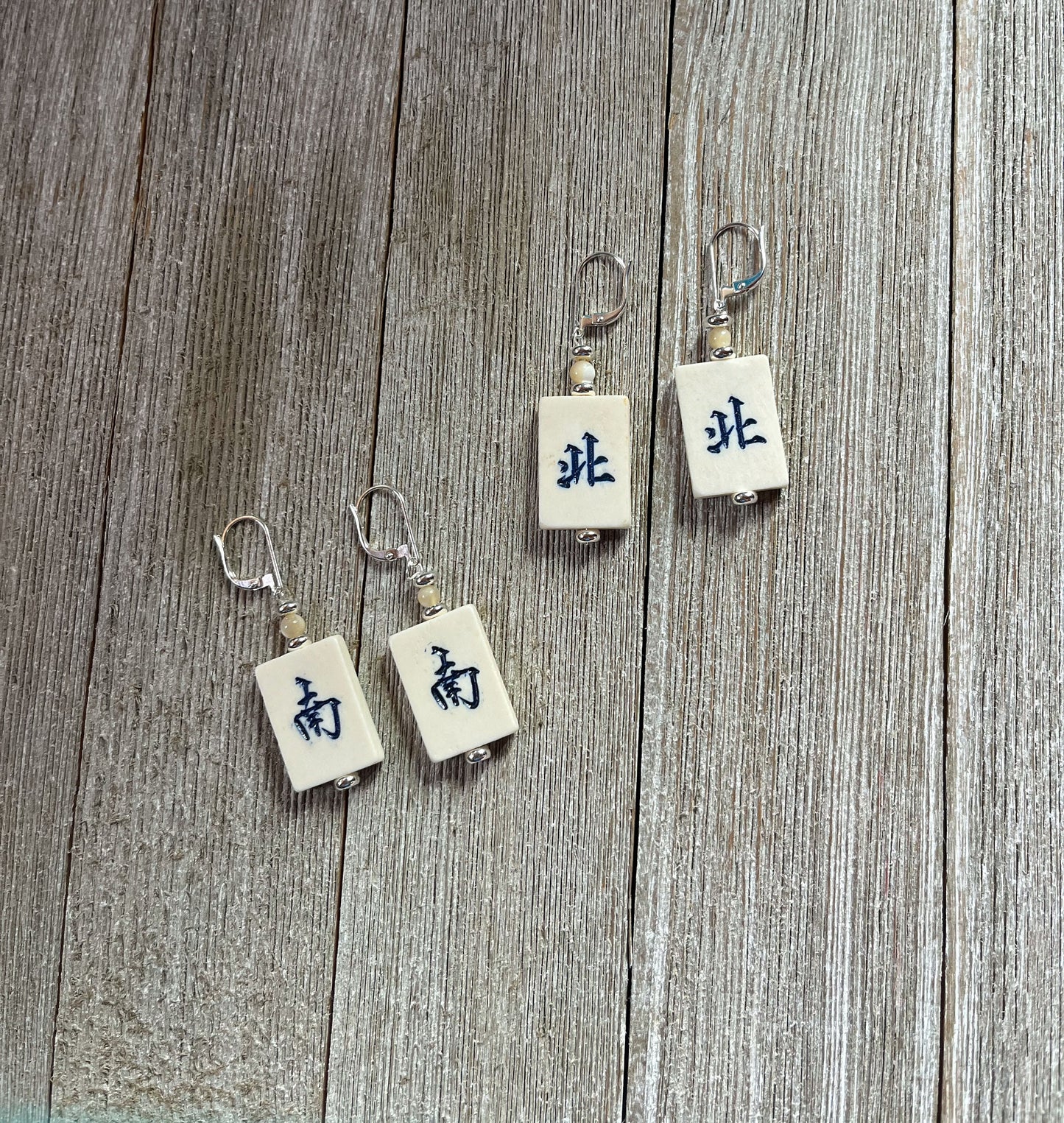 Four Vintage Style Majong Tile Earrings: Handcrafted North/South Winds, each with metal hooks and white tiles featuring black Chinese characters, are displayed on wood. Two earrings share the same wind tile; the other two show different winds.