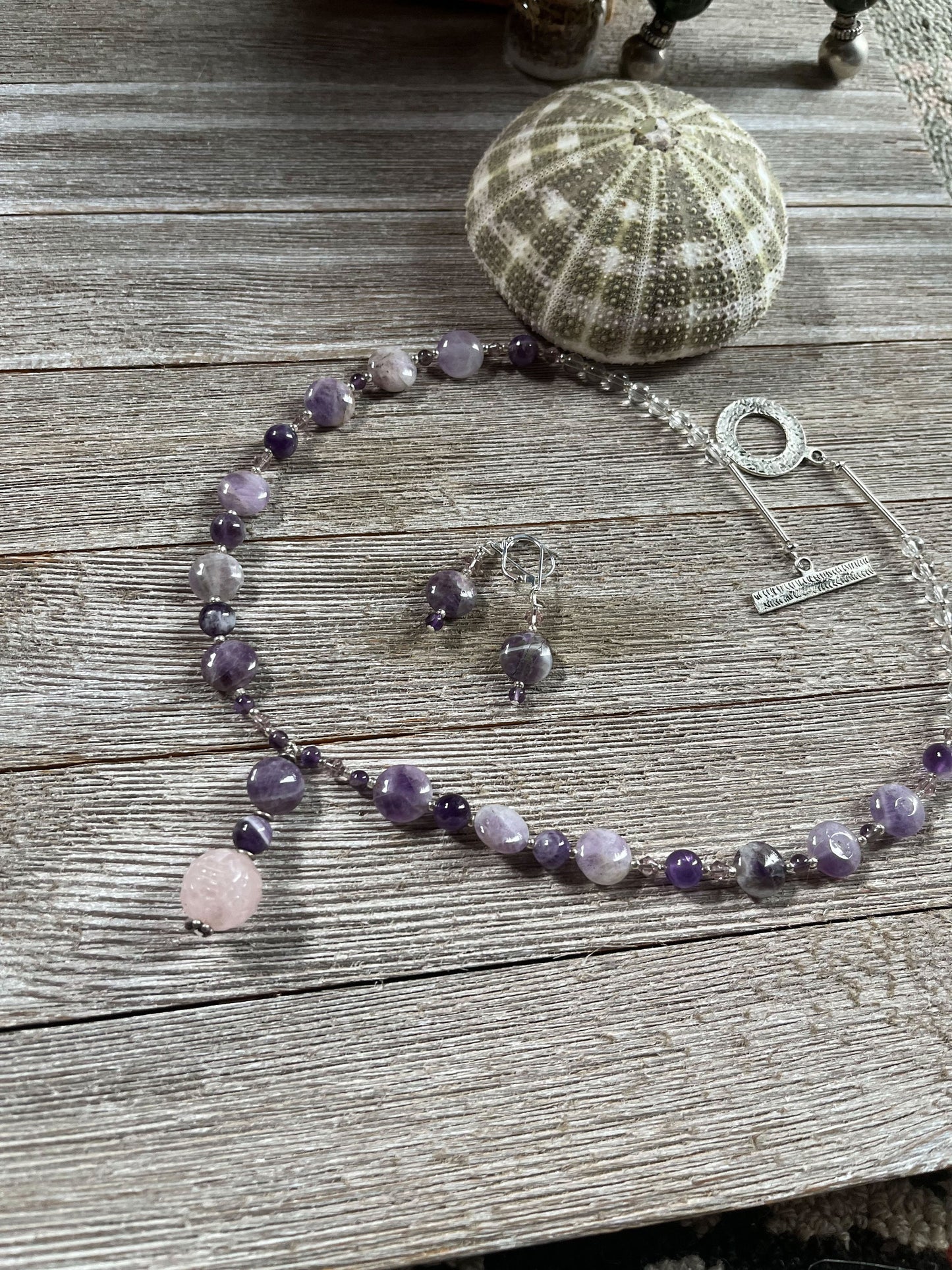 The Artisan Designed Jewelry Set features an amethyst and rose quartz necklace with sterling silver earrings, USA handcrafted, displayed on a rustic wooden surface next to a green sea urchin shell.