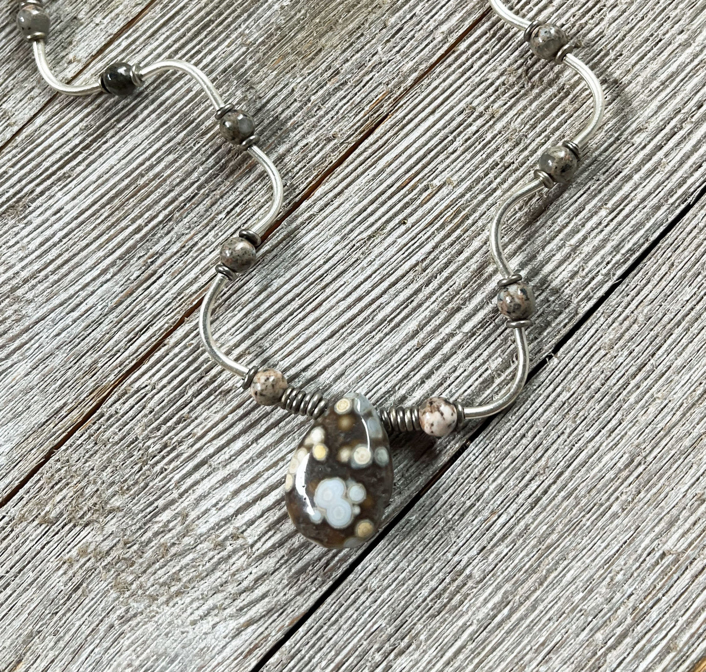 The Ocean Jasper Sterling Silver Jewelry Set features an artisan wave pendant necklace with a wavy design and a sterling silver teardrop pendant adorned with white and yellow flower patterns, displayed on weathered wood.