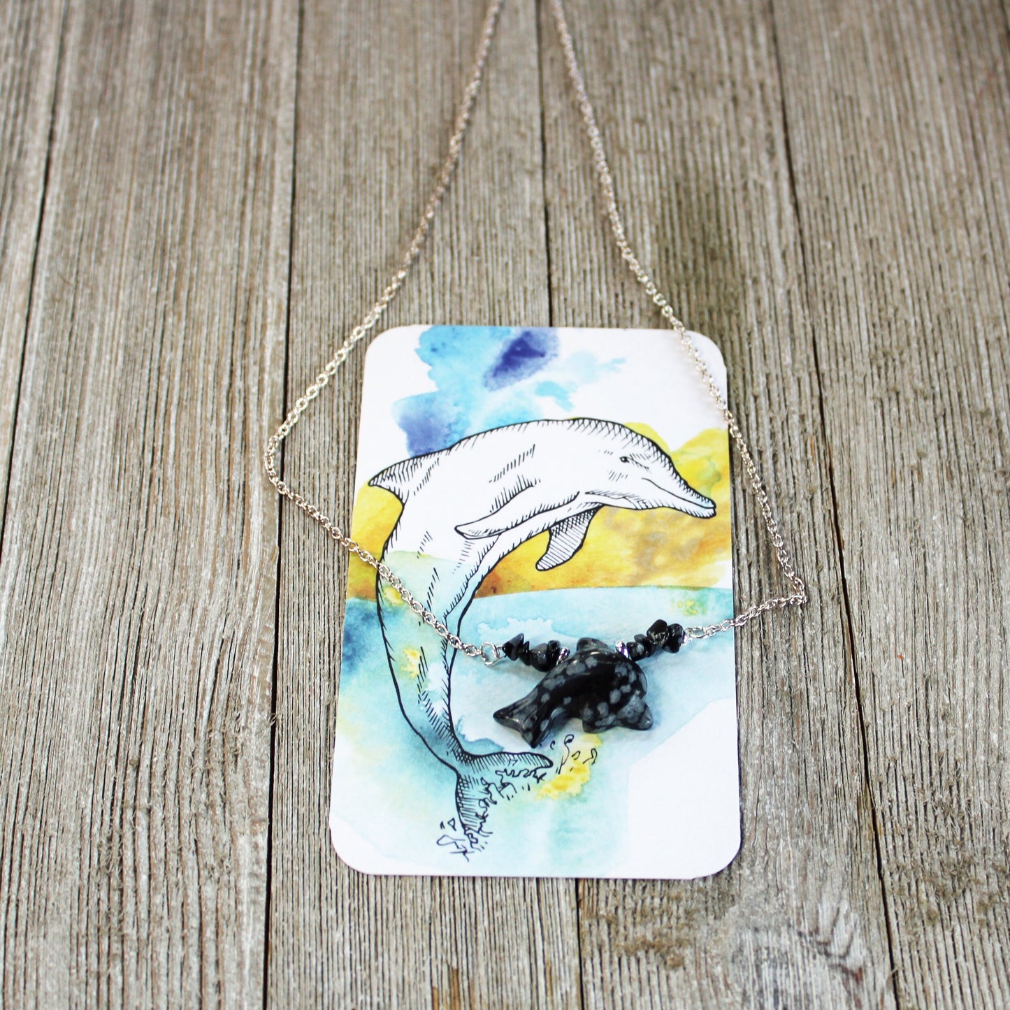 The Handcrafted Snowflake Obsidian Dolphin Necklace: Sea Life Jewelry is shown atop an illustrated card with a dolphin leaping from water, set on rustic wood, showcasing its unique stone beads and ocean-inspired charm.
