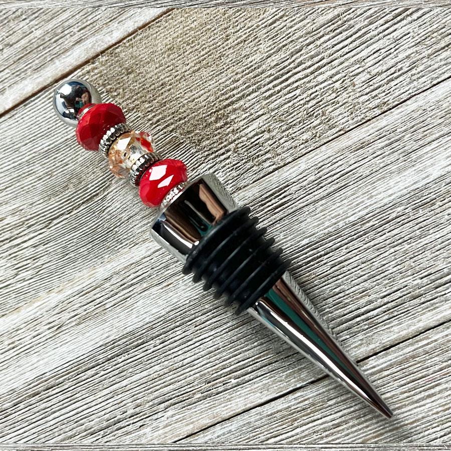 The Handcrafted Red Beaded Wine Stopper – Unique Hostess Gift features shiny silver, gold, and red beads on a metallic handle with black rubber rings—an elegant artisan wine accessory displayed on light wood.