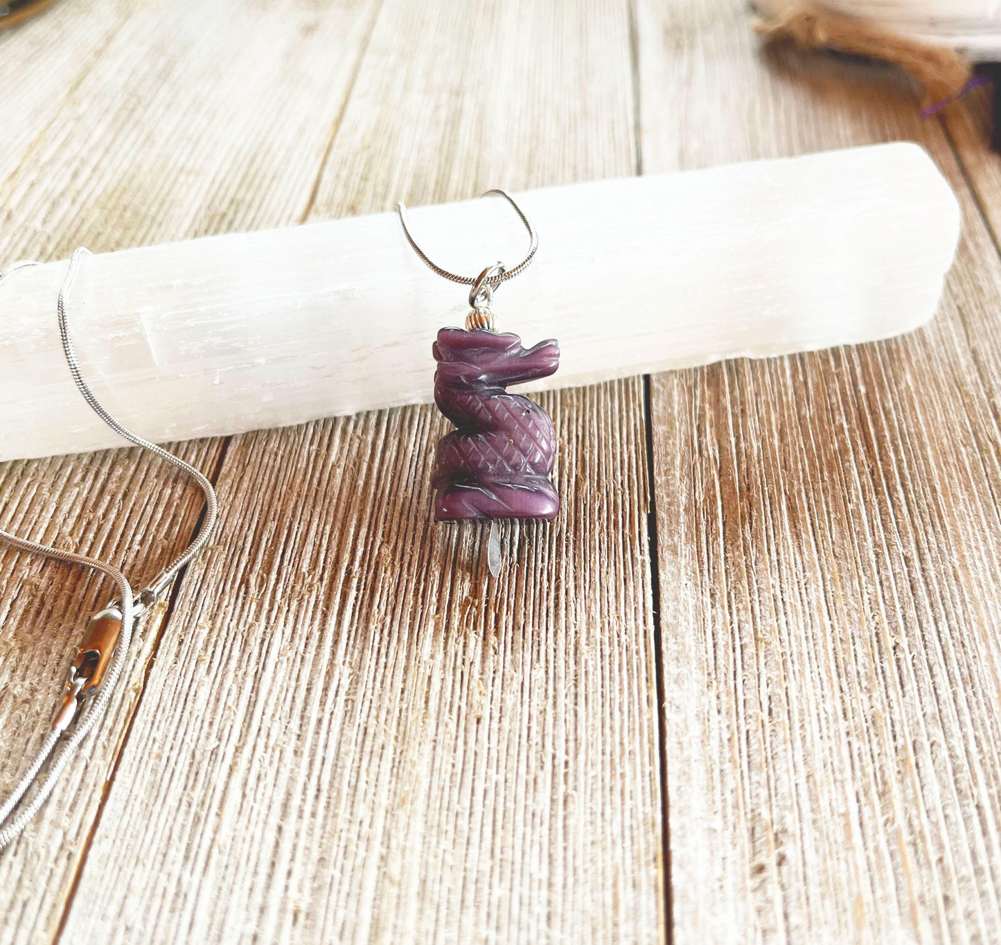 The Purple Dragon Pendant Necklace: Fantasy Glass Jewelry features a spiral-carved purple stone on a silver chain, displayed atop a white crystal wand on a rustic wooden surface.