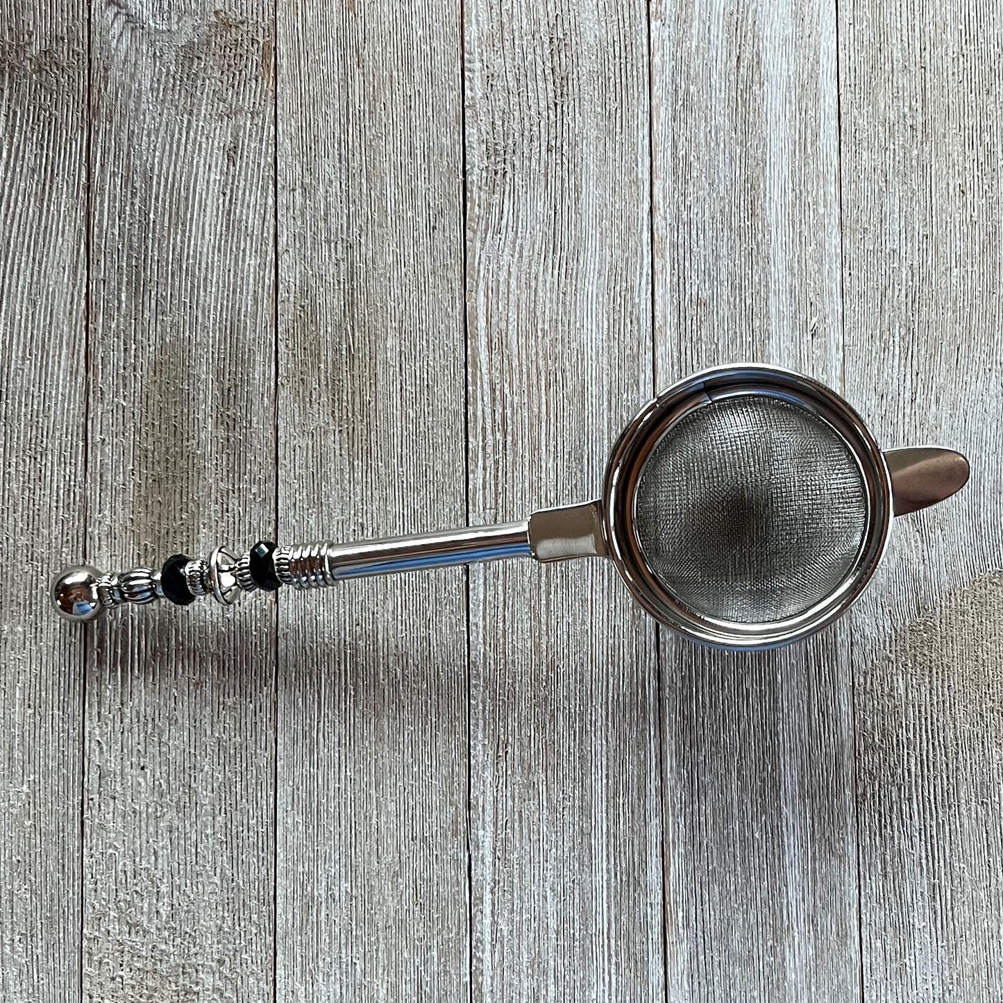The Beaded Tea Strainer – Handcrafted Customizable Handle (9” Length), adorned with black and silver accents, rests elegantly on a light wooden surface—an ideal gift for tea lovers.