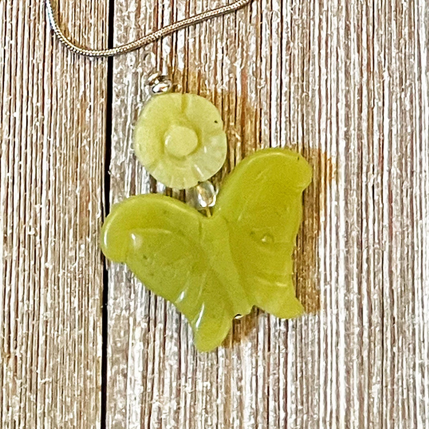 The Carved Jadeite Butterfly Pendant with Flower – Spring Nature Jewelry features a green butterfly pendant on a silver chain, beautifully showcased on textured wood.