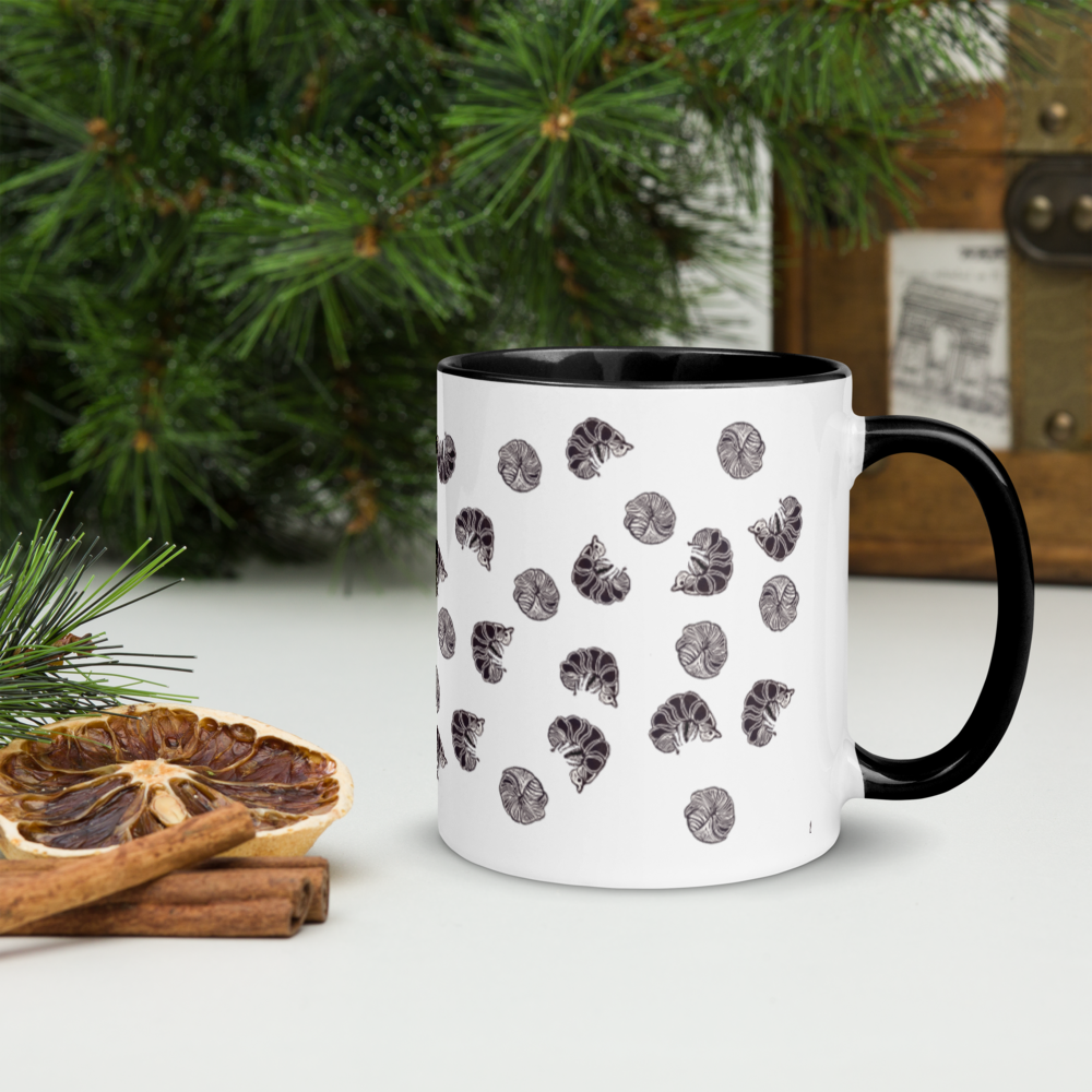 Certainly! Here’s a rewritten version using your product data: The Roly Poly Pill Bug Isopod Mug with Color Inside by Enlivenadorn (Gwynstone) sits on a table near dried citrus slices, cinnamon sticks, pine branches, and a wooden box in the background—a perfect find for any arthropod enthusiast or mug collector who appreciates unique designs.