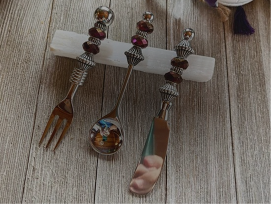 A fork, spoon, and knife with beaded metal handles rest on a white holder atop a wooden surface.