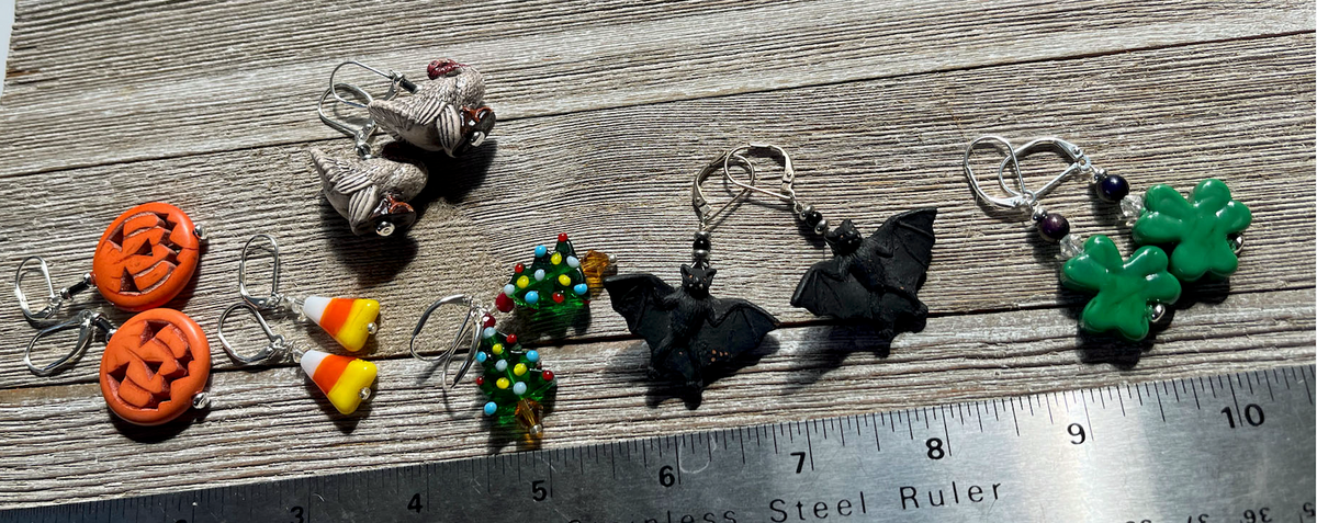 Six pairs of Enlivenadorn by Gwynstone Halloween Theme Earrings—featuring fun designs such as jack o’ lanterns, candy corn, turkeys, Christmas wreaths, bats, and green clovers—are displayed on a wooden surface beside a stainless steel ruler for scale. These earrings are perfect for adding festive flair to any spooky party!.