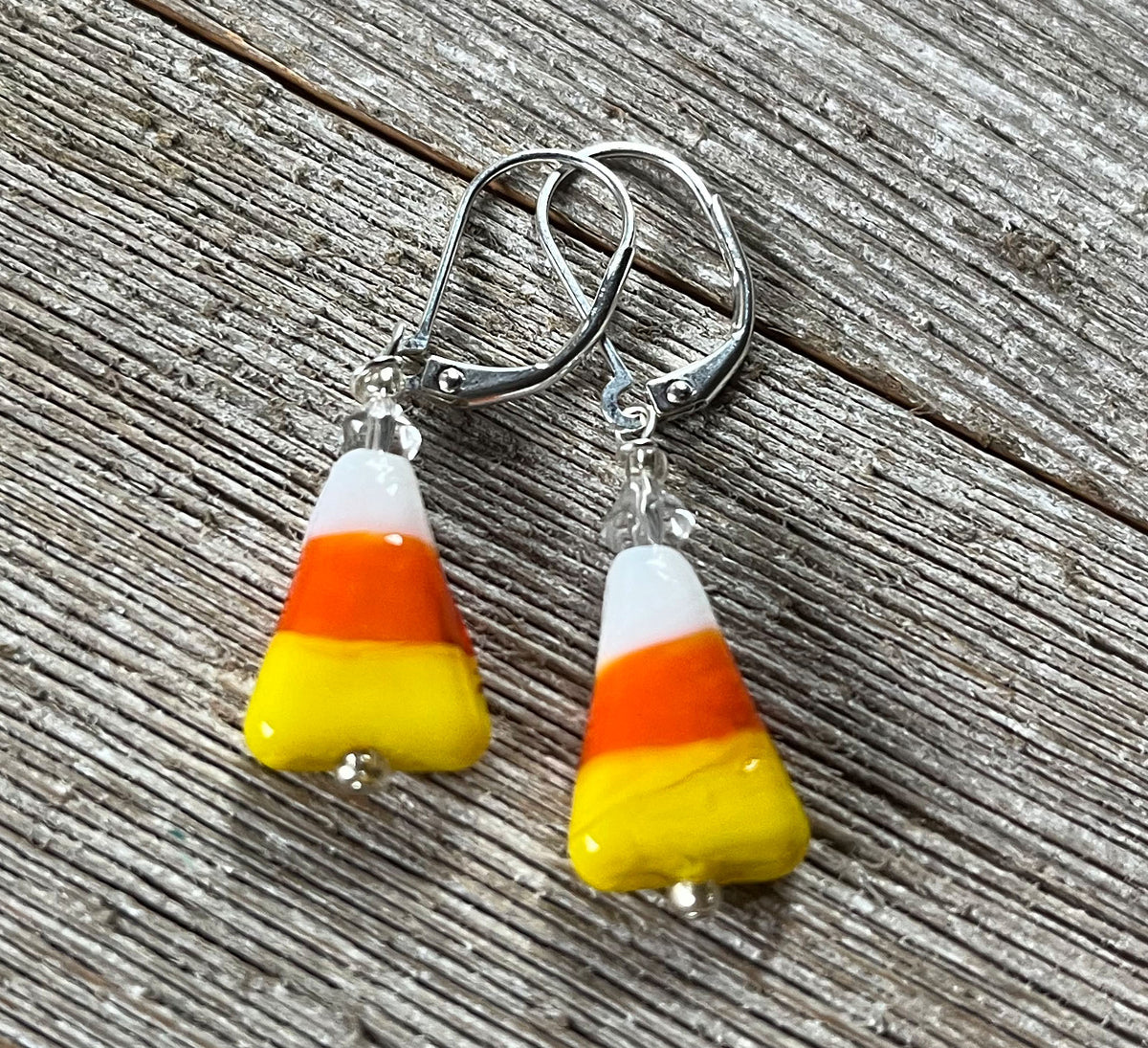 A pair of Halloween Theme Earrings shaped like candy corn, from Enlivenadorn by Gwynstone, featuring white, orange, and yellow stripes with silver hooks, displayed on weathered wood.