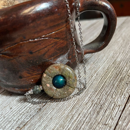The Transform Your Daily Style with the Jasper Mood Bead Necklace - Unique Color-Changing Jewelry by Enlivenadorn by Gwynstone rests against a brown ceramic mug on a rustic wooden surface.