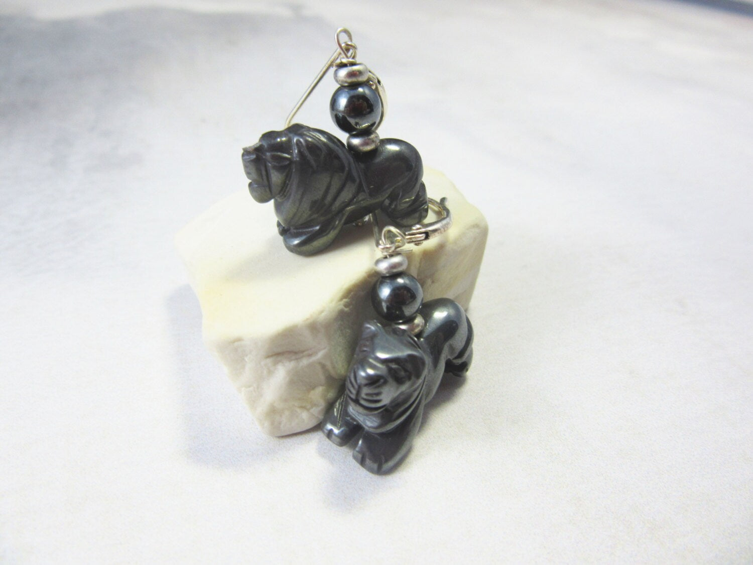 The Lion Hematite Earrings – Animal Themed Grounding Stone Jewelry feature carved black stone lion figures with round beads, showcased on a white, irregular stone surface against a light gray background.