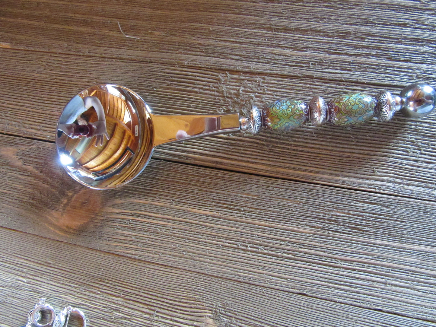 The Artisan Crafted Herb & Bath Salt Spoon Color Changing Mood Bead Scoop, with its vibrant patterns, silver accents, and decorative handle, rests on a wooden surface. Its round bowl shines beautifully, reflecting the surroundings.