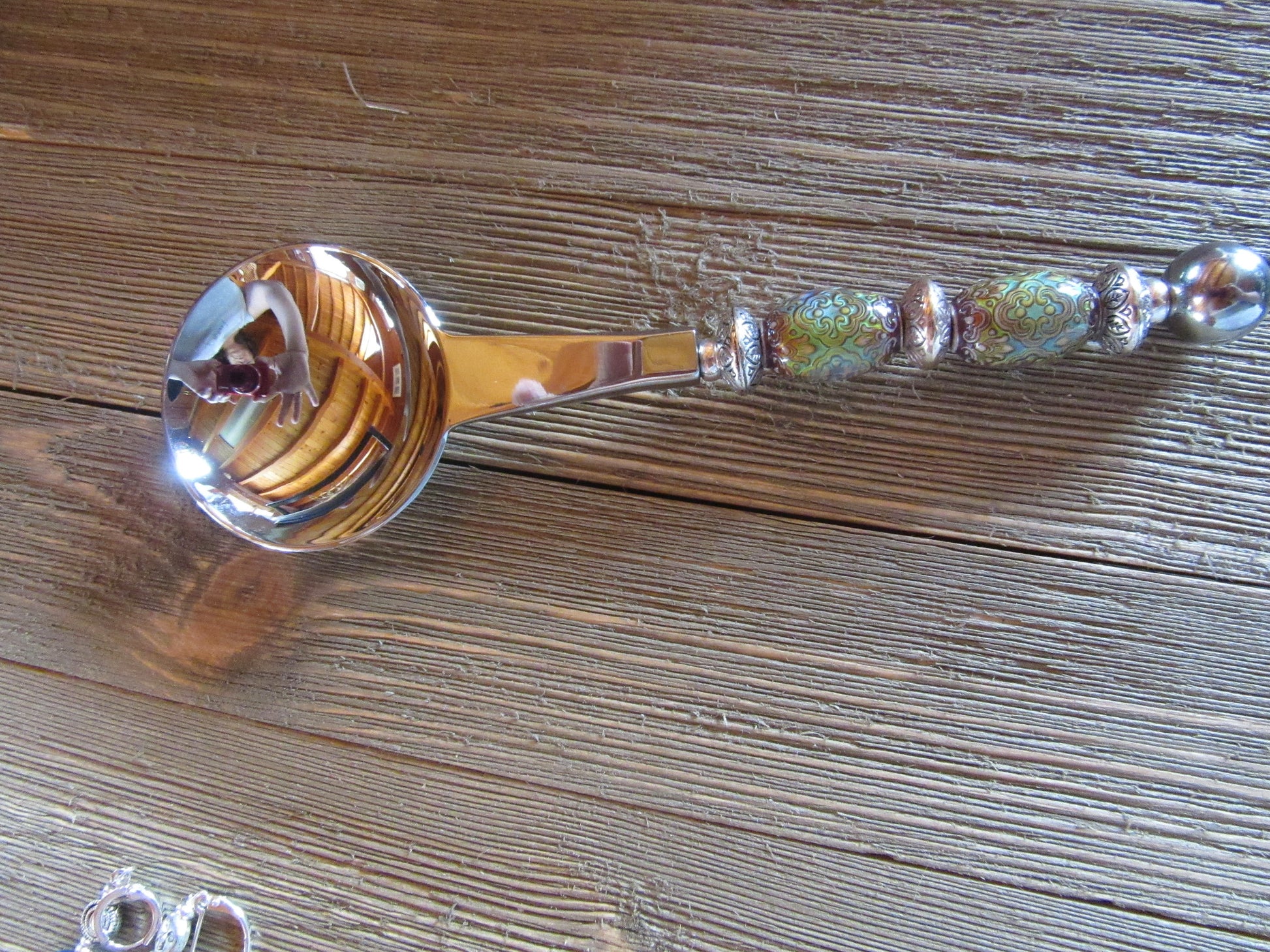The Artisan Crafted Herb & Bath Salt Spoon Color Changing Mood Bead Scoop, with its vibrant patterns, silver accents, and decorative handle, rests on a wooden surface. Its round bowl shines beautifully, reflecting the surroundings.
