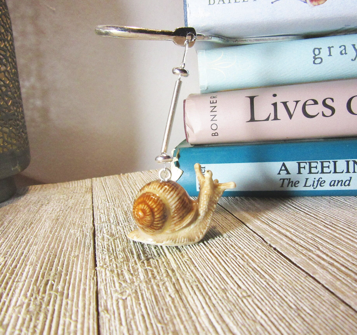 A charming porcelain snail bookmark with a silver metal clip sits on a wooden surface by pastel books—an ideal handcrafted gift for book lovers, perfect beside your favorite reads or paired with another whimsical reading accessory.