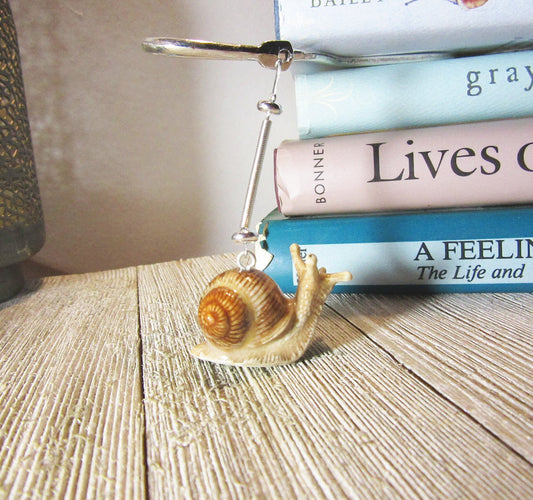 A charming porcelain snail bookmark with a silver metal clip sits on a wooden surface by pastel books—an ideal handcrafted gift for book lovers, perfect beside your favorite reads or paired with another whimsical reading accessory.