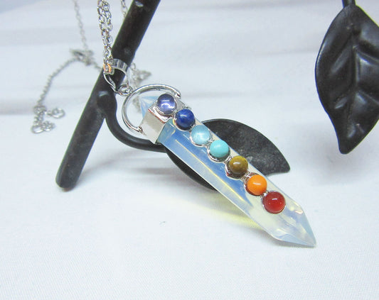 The Handcrafted Opalite Wand Pendant: Chakra Stone Necklace features seven colorful chakra stones on a silver chain, elegantly displayed over a black metal leaf—a distinctive piece of handmade jewelry.