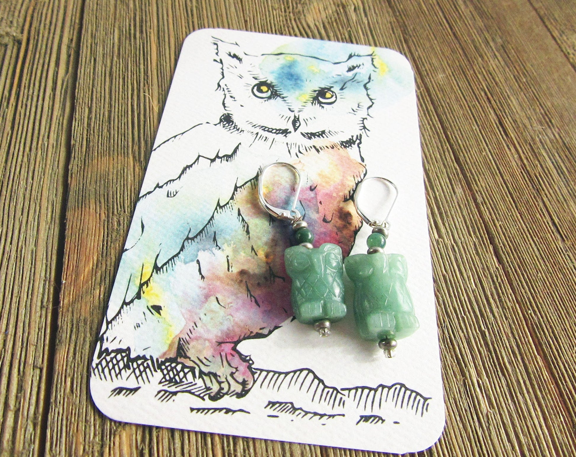 Handcrafted Owl Earrings: Natural Green Stone, Sterling Silver-Filled, are displayed on a card with a watercolor and ink owl illustration, all set atop a rustic wooden surface.