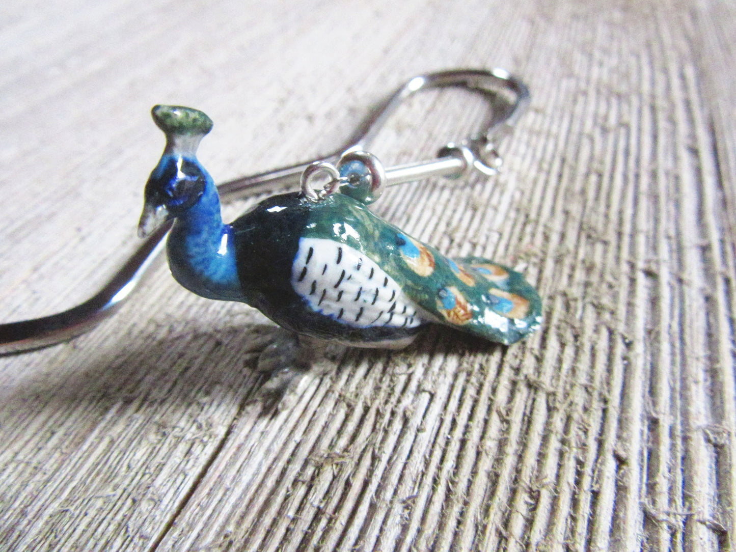 A close-up of the Hand-crafted Porcelain Peacock Bookmark, a unique reader gift with blue, green, and orange details on a metallic keychain, shown resting on a textured wooden surface.