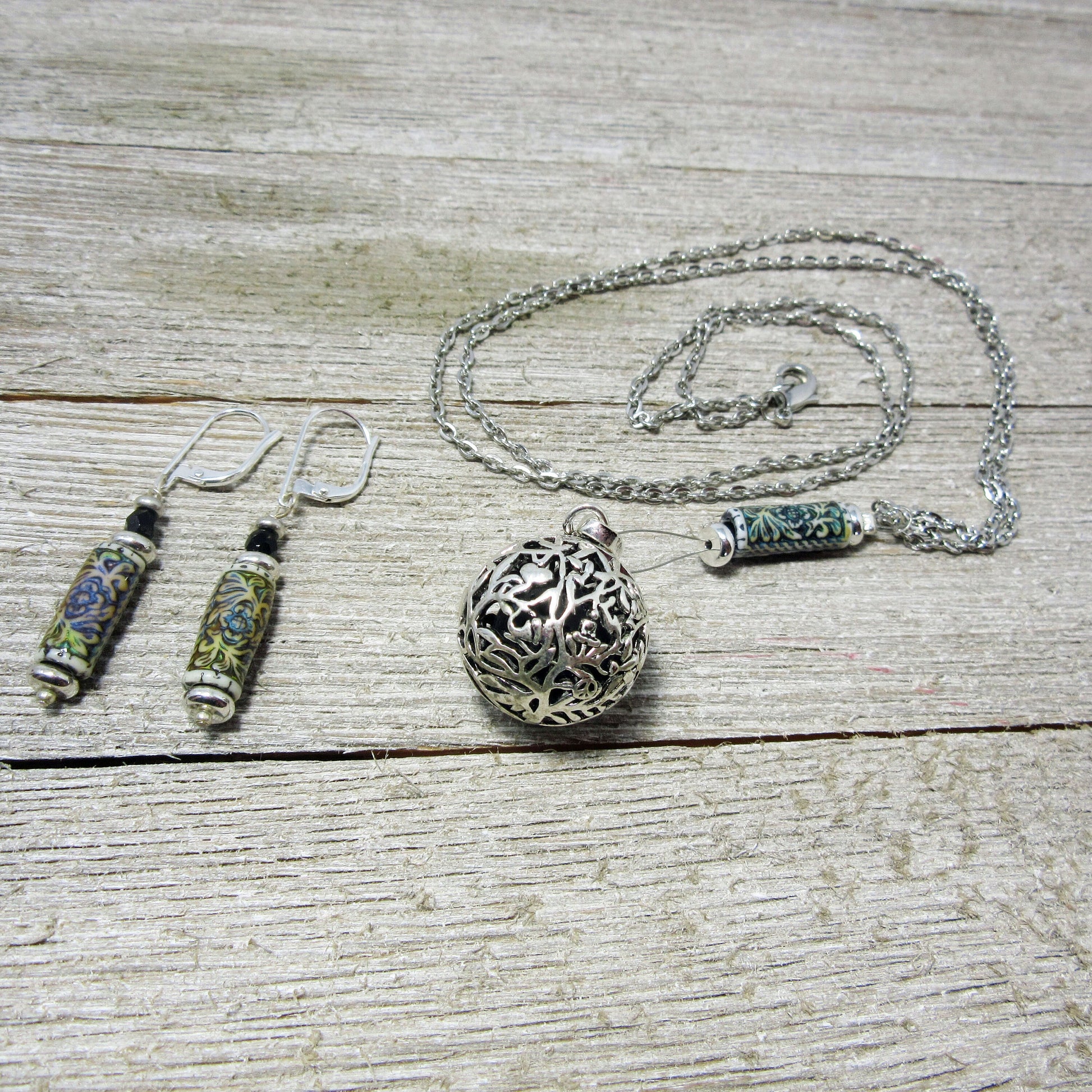 The Floral Diffuser Necklace Earring Set: Color Changing Aromatherapy Jewelry features intricate metalwork with floral designs in green and blue, displayed on a weathered wood surface. Includes dangling earrings and a filigree pendant necklace.