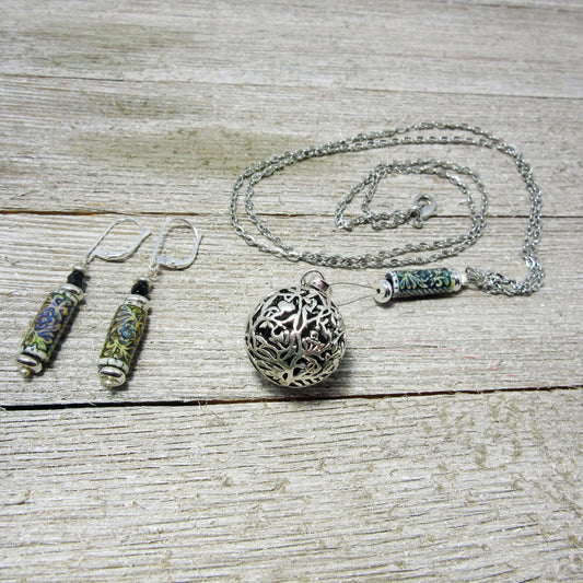 The Floral Diffuser Necklace Earring Set: Color Changing Aromatherapy Jewelry features intricate metalwork with floral designs in green and blue, displayed on a weathered wood surface. Includes dangling earrings and a filigree pendant necklace.