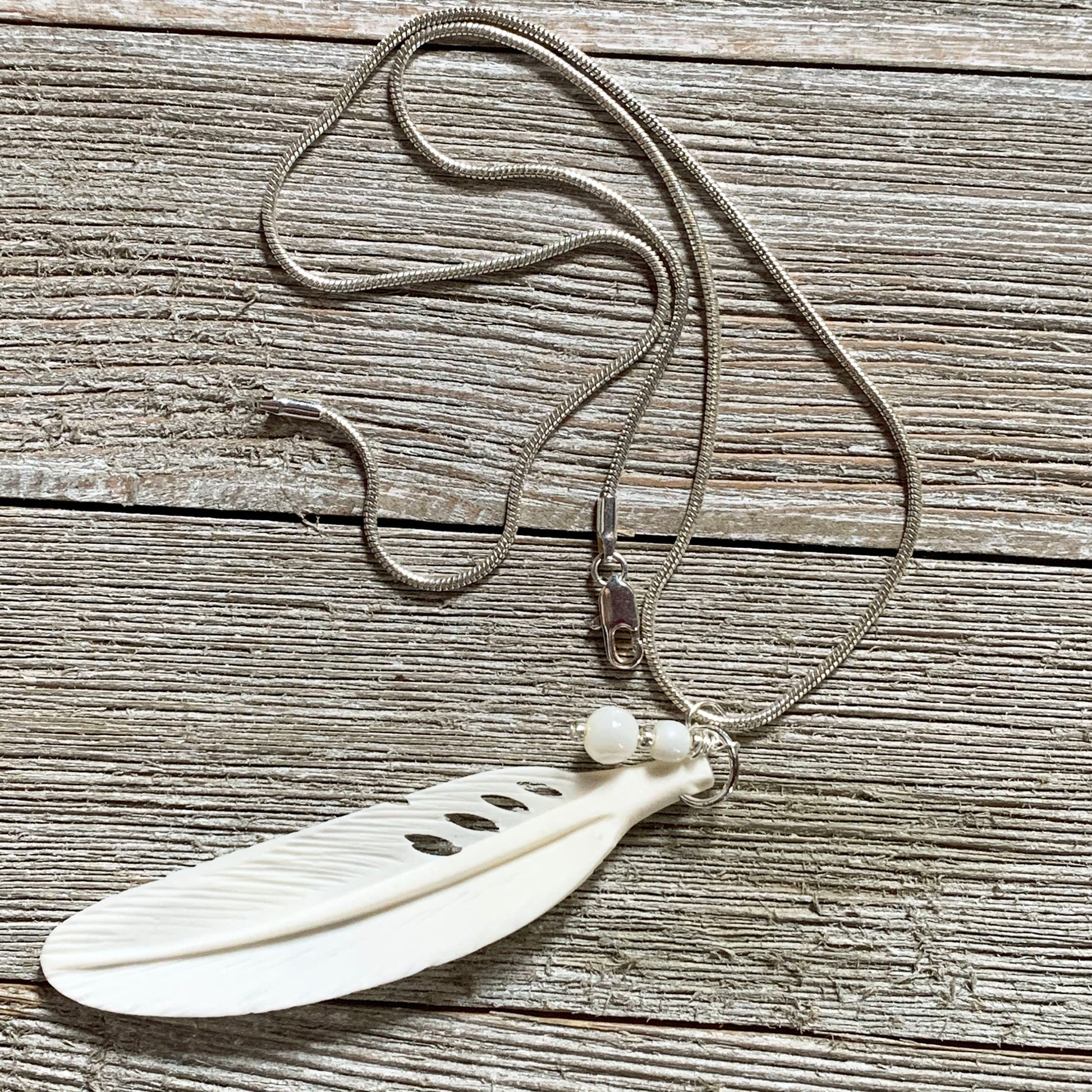 The White Feather Necklace features an angel sign carved bone and mother of pearl pendant, displayed on a rustic wooden surface—ideal for those seeking meaningful spiritual jewelry.