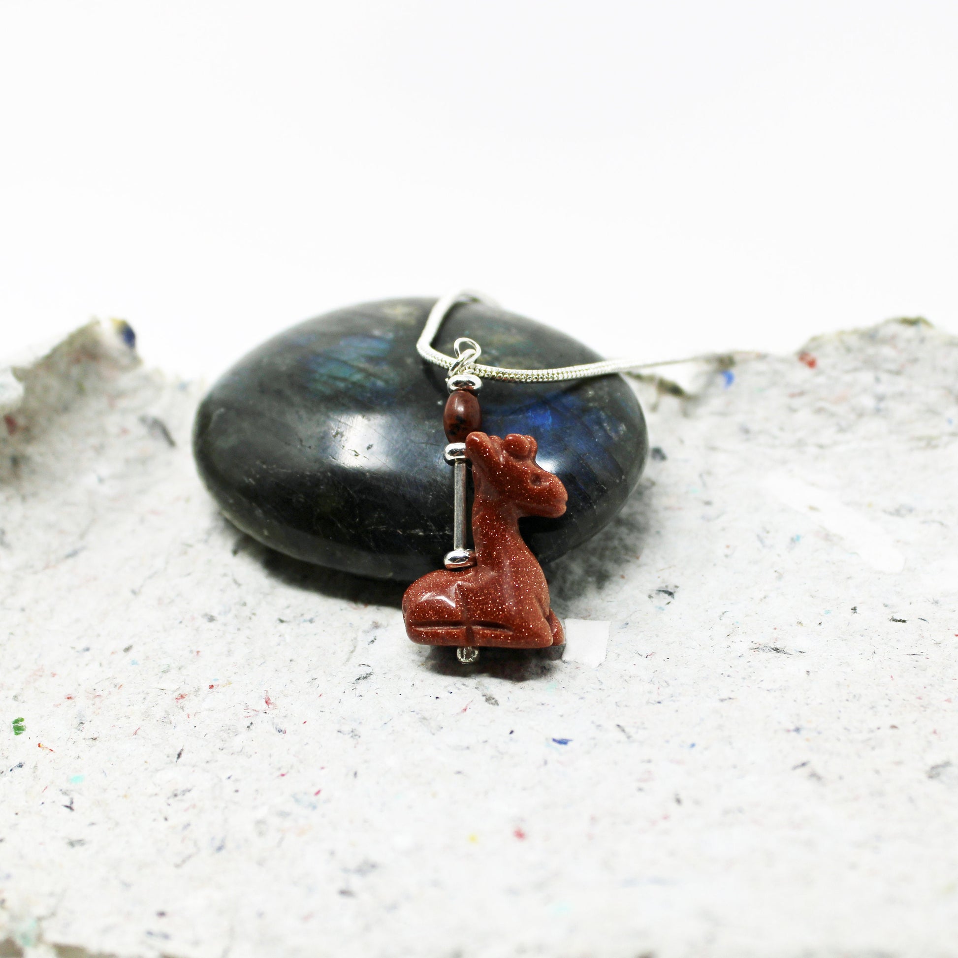 The Handcrafted Goldstone Giraffe Charm Pendant, designed for animal lovers, features a silver giraffe charm and goldstone pendant displayed on a textured gray surface with a polished dark stone in the background.
