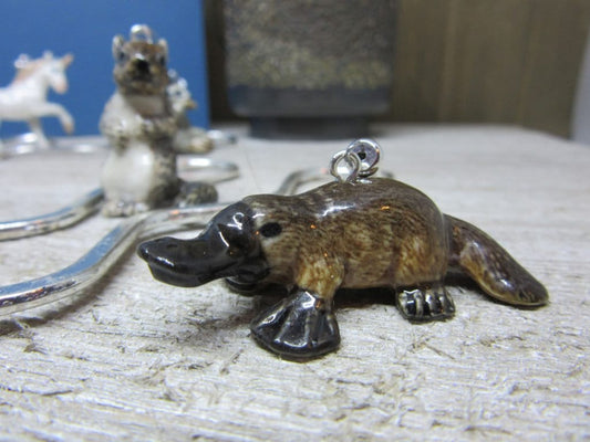 The Exotic Porcelain Platypus Figurine Bookmark for Book Reader Gift, featuring a handy metal loop, rests on wood as an ideal whimsical page marker. A blurred ceramic animal and silver object enhance the scene's charm in the background.