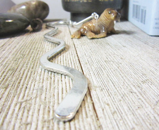 A close-up of the Porcelain Walrus Bookmark, a handmade metal reader gift, on a wooden surface with a small walrus figurine and round stones in the background—perfect for book lovers.