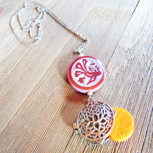 The Dragon & Peacock Carnelian Essential Oil Diffuser Necklace: Lotus Zen Jewelry features a silver chain, a red carnelian pendant with bird detail, and a silver filigree locket with yellow felt, displayed on wood.