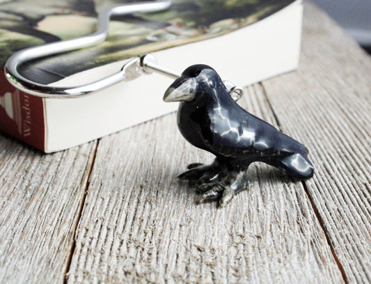 A hand-painted raven figurine bookmark rests on a wooden surface near the edge of an open book, alongside a unique metal book lover gift shaped like a hook.
