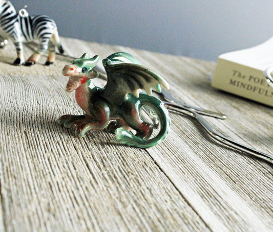 A small green dragon figurine sits on wood. In the blurred background, a zebra figurine, a book, and the Fantasy Green Dragon Bookmark—porcelain and metal, perfect for fantasy readers—can be seen.