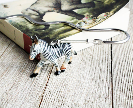 A zebra-shaped keychain hook sits on wood beside a book with an animal cover, making it a fun reading gift or a perfect match for the Porcelain Zebra Bookmark – Animal Lover Gift for Readers, Book Fans.