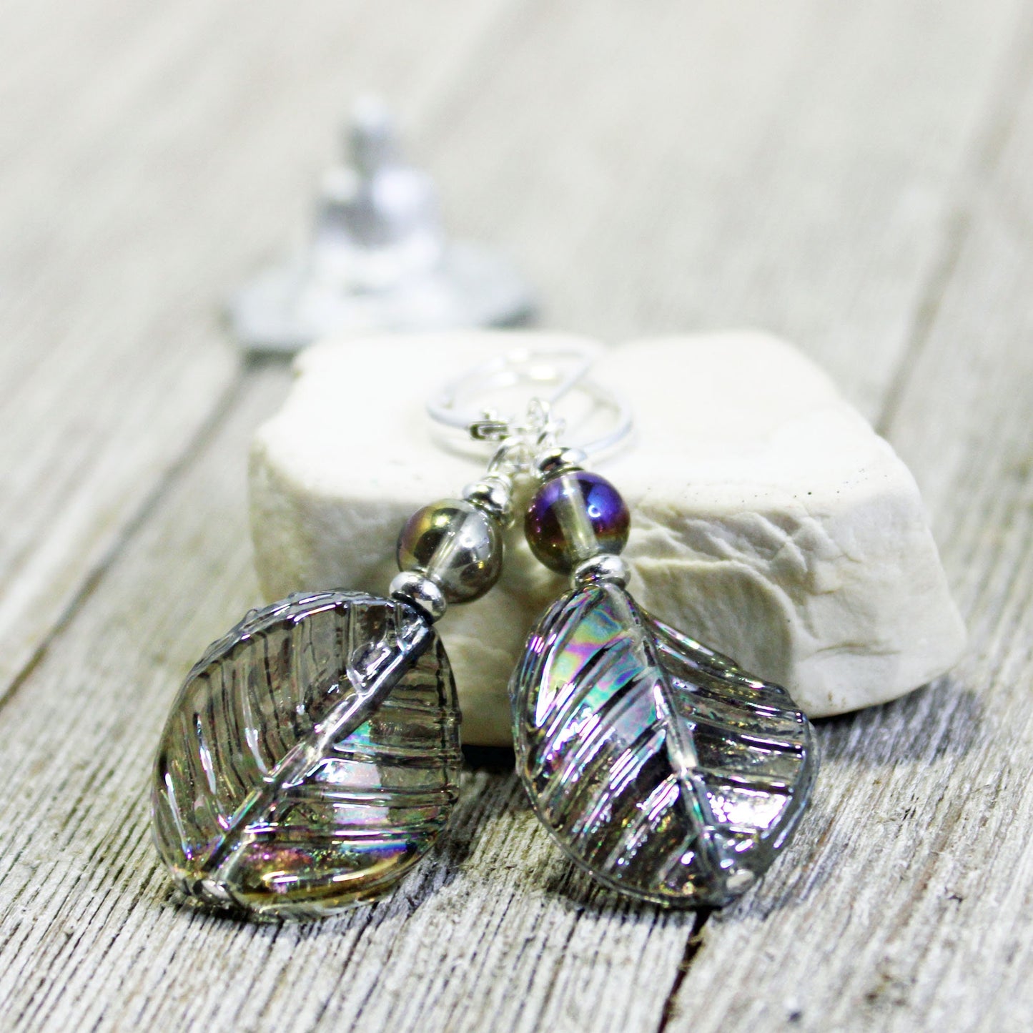 The Shimmering Crystal Leaf Earrings - Nature Lover's AB Finish Gift feature iridescent leaf shapes with metallic accents, displayed on a white stone atop rustic wood—perfect nature-inspired jewelry for any occasion.