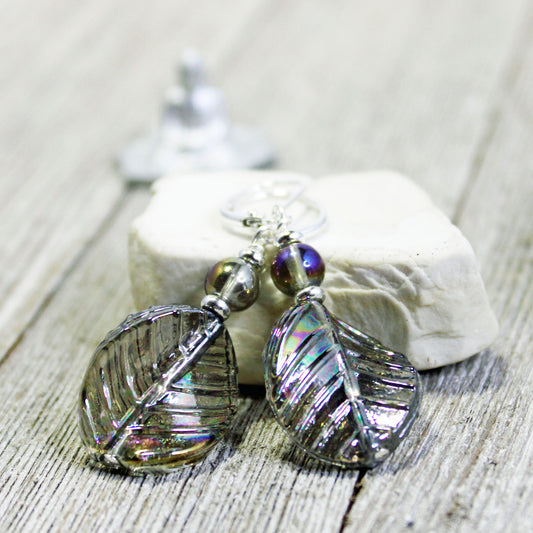 The Shimmering Crystal Leaf Earrings - Nature Lover's AB Finish Gift feature iridescent leaf shapes with metallic accents, displayed on a white stone atop rustic wood—perfect nature-inspired jewelry for any occasion.