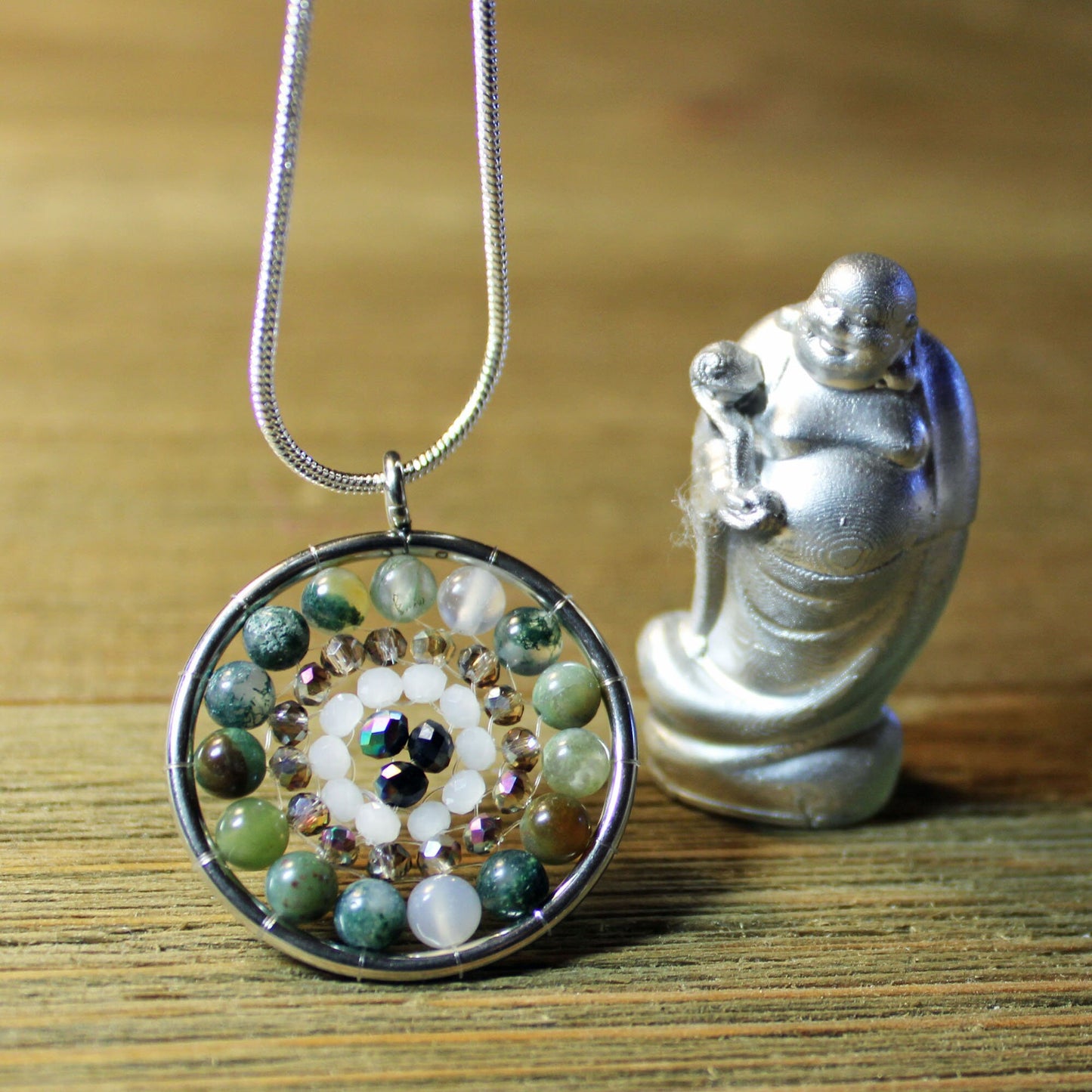 A Zen Jasper Pendant Necklace: Aurora Borealis Crystals, Handcrafted Jewelry featuring green, white, and black stones rests on wood beside a silver Buddha figurine holding a staff.