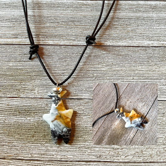 Displayed on a wooden surface, the Shark Pendant Necklace features a handcrafted amazonite shark-shaped pendant in black, white, and tan on a black cord. An inset offers a close-up of this unique ocean theme jewelry piece.
