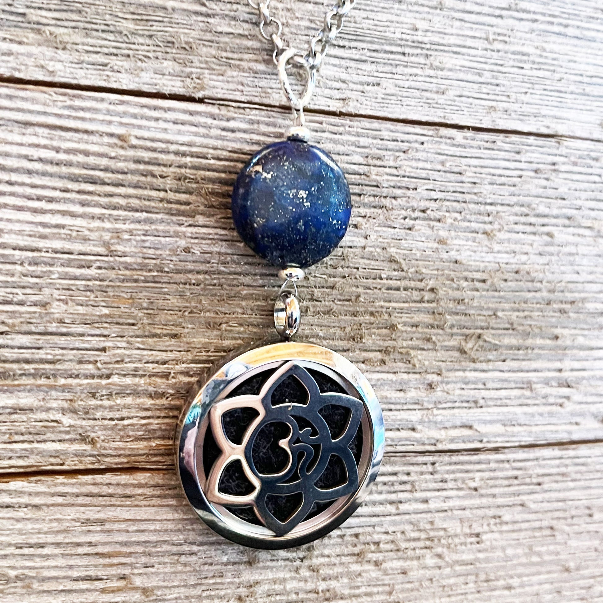 The Lapis Lazuli Aromatherapy Necklace & Sterling Silver Earrings set features a round lapis lazuli stone above an oval lotus locket, shown on a textured wooden background—a spiritual and elegant jewelry set.