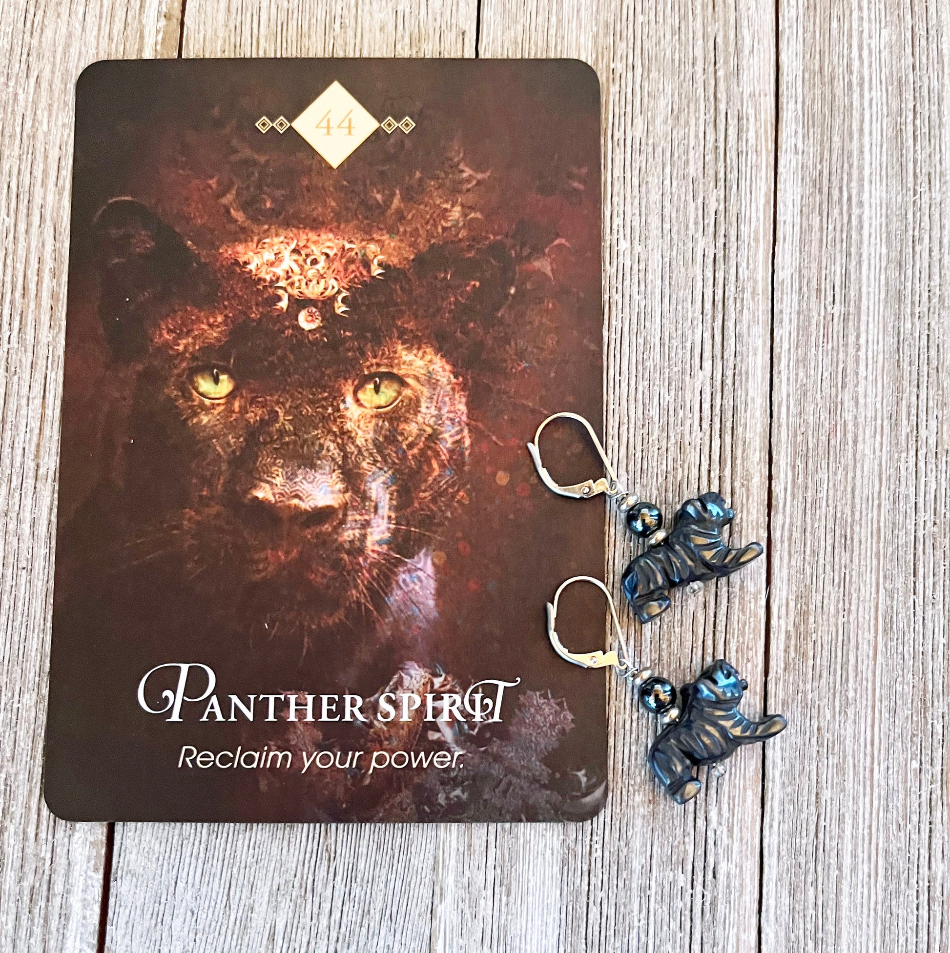 A card showing a black panther, "Panther Spirit," "Reclaim your power," and the number 44 rests on wood beside Enchanting Panther Hematite Earrings—unique animal jewelry perfect for big cat lovers with grounding blue accents.