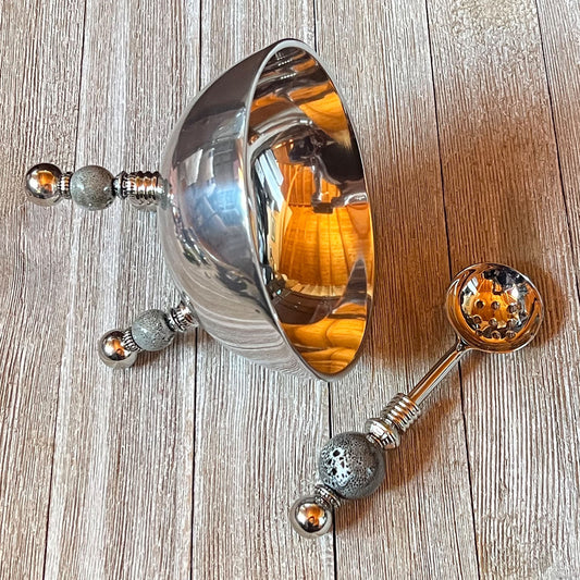 The Stainless Steel Bowl Set features gray ceramic legs and a matching spoon, both with intricate handle designs and reflective surfaces. Ideal as an altar bowl set or spiritual jewelry organizer, they rest elegantly on any wooden surface.