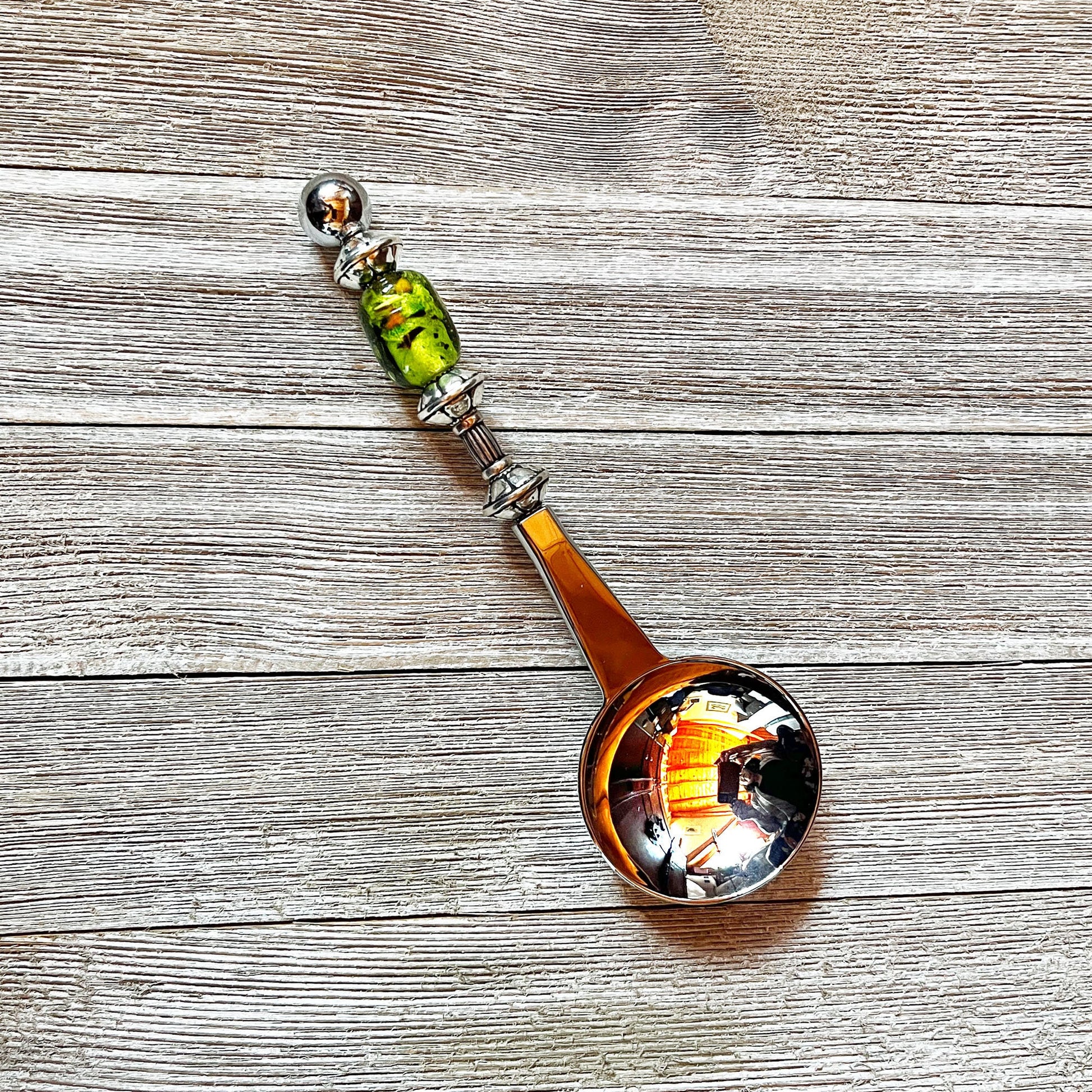 The Art Glass Beaded Handle Coffee Spoon, a stainless steel 1 Tbsp measure with a green art glass bead on its handle, rests on a textured wooden surface. The spoon’s bowl reflects colorful indoor lights and surroundings.