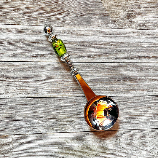 The Art Glass Beaded Handle Coffee Spoon, a stainless steel 1 Tbsp measure with a green art glass bead on its handle, rests on a textured wooden surface. The spoon’s bowl reflects colorful indoor lights and surroundings.