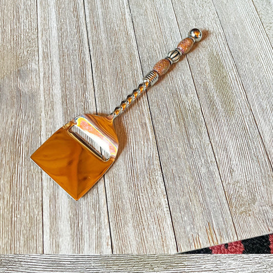 The Color Changing Beaded Cheese Slicer - Stainless Steel Party Tableware, featuring a decorative handle, sits on a light wooden surface that showcases its unique beaded design and the wood's visible grain patterns.