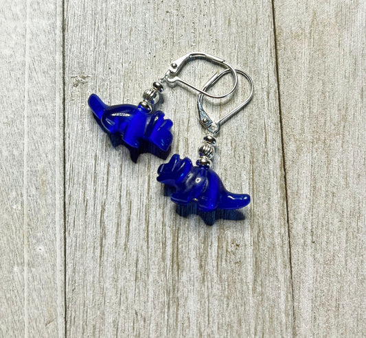 A pair of Triceratops Dinosaur Earrings—handcrafted with blue optic cat eye style charms on silver hooks—displayed on a light wooden surface.