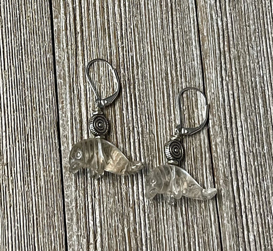 Manatee Earrings: Carved clear quartz charms with a decorative spiral hang from sterling silver lever backs, displayed on a wooden surface.