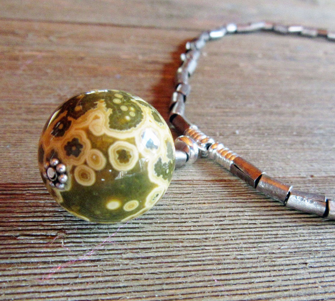A close-up of the Ocean Jasper Atlantis Stone Orb Pendant Necklace, featuring green and yellow patterns in a floral design and metallic cylindrical beads, showcases its artisan-made minimalist style on a wooden surface.