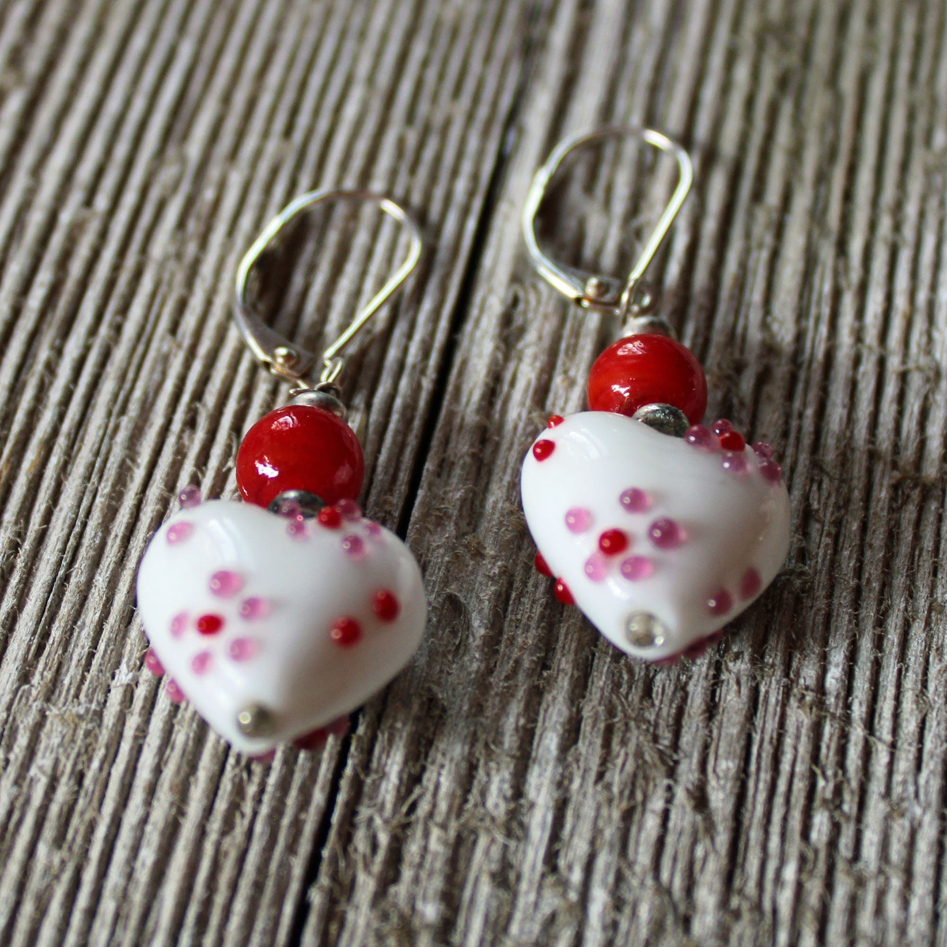 A pair of Lampwork Glass Heart Dangle Earrings—red and white lever back jewelry with heart-shaped beads—displayed on a wooden surface.